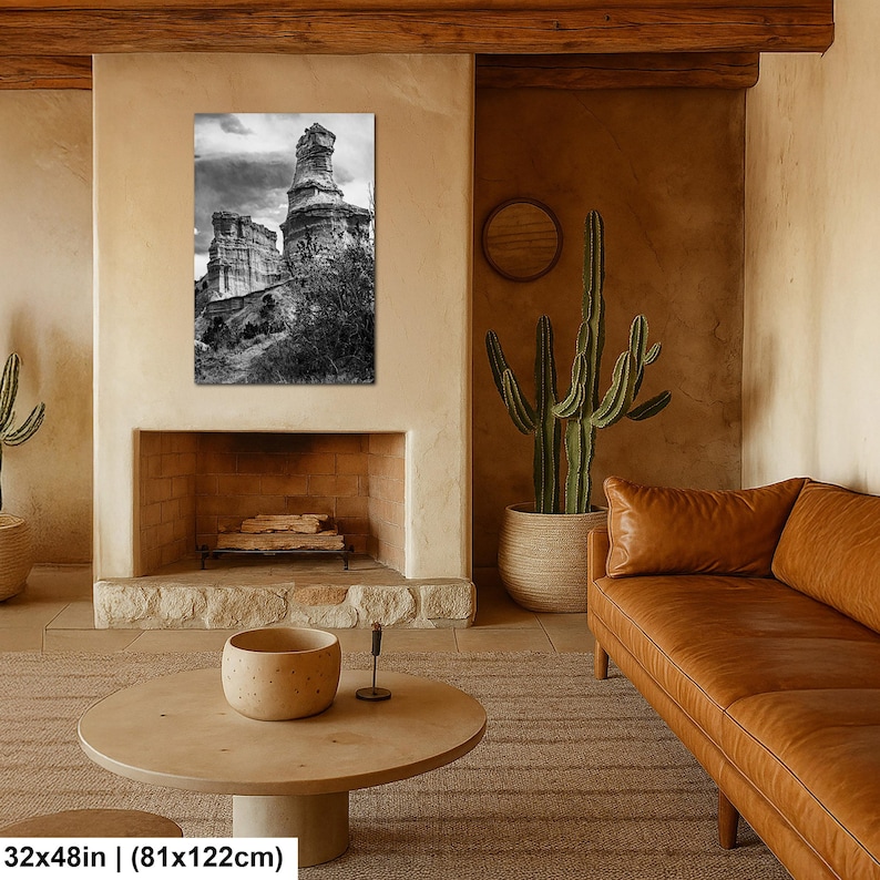 May include: A black and white wall art print depicting a tall, weathered rock formation under a cloudy sky. The rectangular print measures 32 inches by 122 inches and is displayed above a fireplace in a room with a brown leather sofa and cactus.