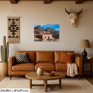 May include: A rectangular wall art print measuring 24 by 36 inches, depicting a tan adobe structure nestled against rugged desert mountains under a bright blue sky. The print is displayed on a beige wall above a brown leather sofa and wooden coffee table.