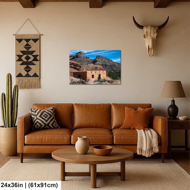 May include: A rectangular wall art print measuring 24 by 36 inches, depicting a tan adobe structure nestled against rugged desert mountains under a bright blue sky. The print is displayed on a beige wall above a brown leather sofa and wooden coffee table.
