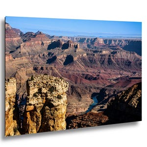 May include: A landscape photograph of the Grand Canyon featuring layered red and brown rock formations, deep shadows, and a winding blue river visible at the base. The scene is illuminated by bright daylight under a clear blue sky.