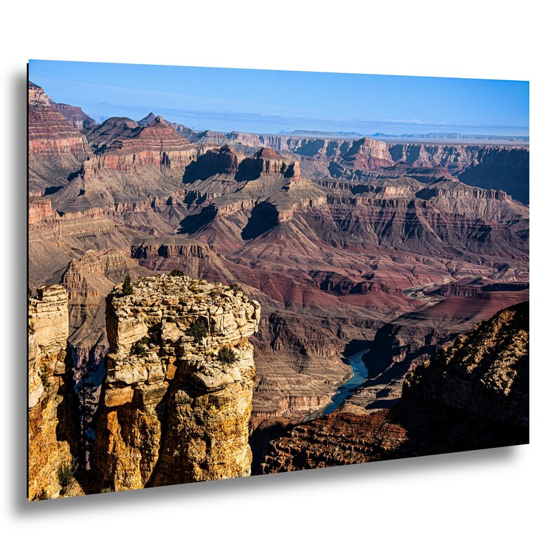 May include: A landscape photograph of the Grand Canyon featuring layered red and brown rock formations, deep shadows, and a winding blue river visible at the base. The scene is illuminated by bright daylight under a clear blue sky.