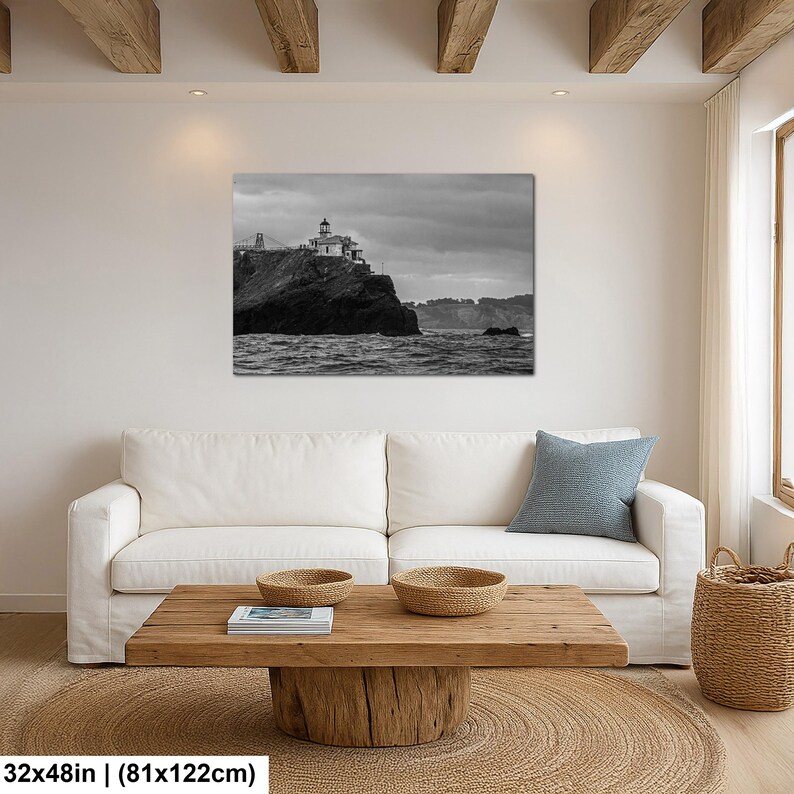 May include: A black and white landscape wall art print featuring a lighthouse perched on a rocky cliff overlooking the ocean under a cloudy sky. The artwork measures 32 inches by 48 inches and is displayed above a white sofa in a living room.