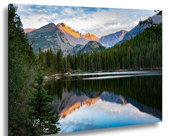 Puede incluir: Una fotografía de paisaje que muestra un lago tranquilo que refleja picos montañosos irregulares iluminados por la luz dorada del sol. Un denso bosque de pinos de color verde oscuro bordea la orilla bajo un cielo con nubes suaves.