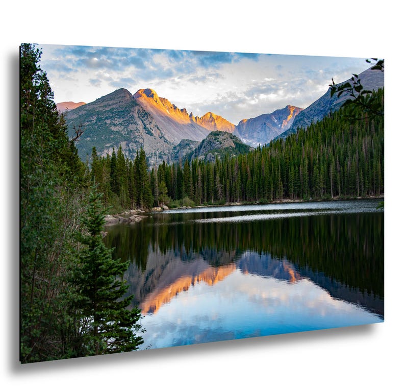 Bear Lake in Rocky Mountain National Park. A landscape photograph showing a calm lake reflecting jagged mountain peaks illuminated by golden sunlight. A dense forest of dark green pine trees lines the shore, while the sky above features soft clouds and blue tones.