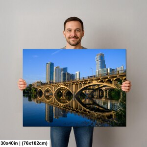 May include: A rectangular wall art print measuring 30 by 40 inches, featuring a scenic landscape photograph of a concrete arched bridge spanning a calm river, with a modern city skyline of skyscrapers under a clear blue sky in the background.
