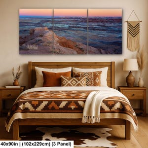 May include: A three-panel wall art set depicting a desert landscape at sunset with orange, blue, and purple hues. The artwork is displayed above a wooden bed frame featuring a brown and cream patterned blanket, pillows, and a cowhide rug.