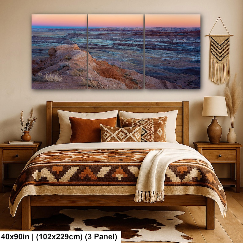 May include: A three-panel wall art set depicting a desert landscape at sunset with orange, blue, and purple hues. The artwork is displayed above a wooden bed frame featuring a brown and cream patterned blanket, pillows, and a cowhide rug.