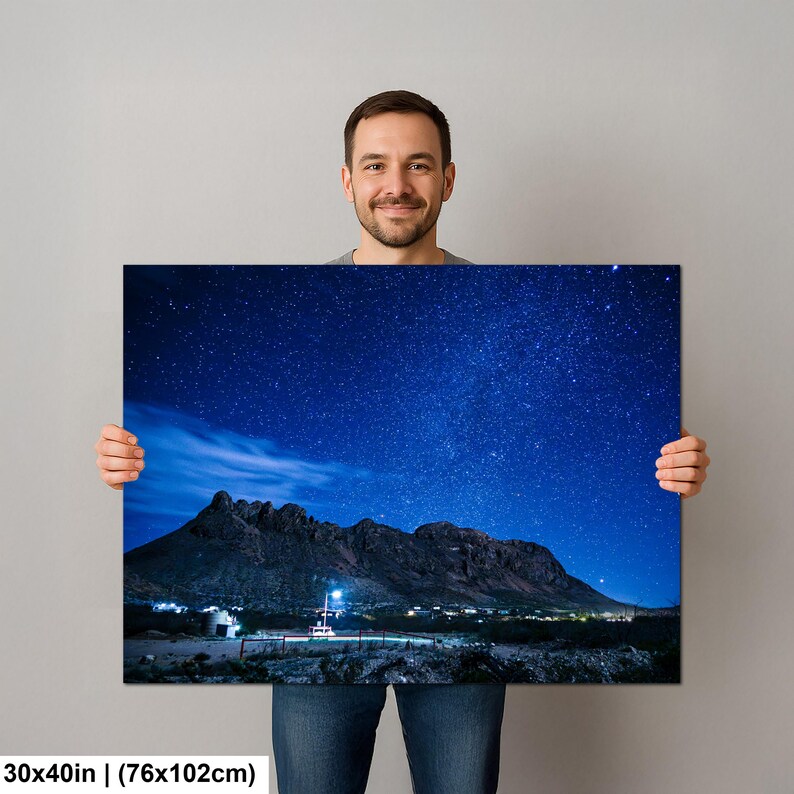 Puede incluir: Una impresi&oacute;n de arte mural rectangular de 76 por 102 cm, que muestra un paisaje nocturno de una cordillera oscura bajo un cielo estrellado azul profundo. La escena incluye una peque&ntilde;a estructura iluminada en la base de las monta&ntilde;as.