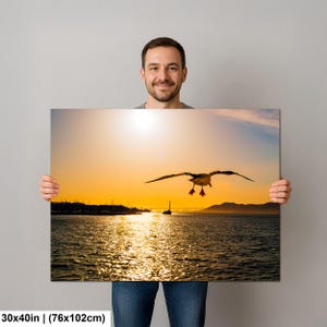 May include: A rectangular wall art print measuring 30x40 inches featuring a sunset over a body of water. A seagull is captured in flight in the center against a bright orange sky. A distant bridge silhouette is visible on the horizon line.