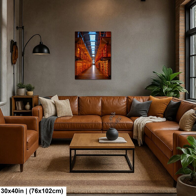 May include: A rectangular wall art print measuring 30x40 inches, depicting a long, narrow prison corridor with orange-lit cell blocks on both sides, a central walkway, and a blue-tinted ceiling structure, displayed above a brown leather sofa.