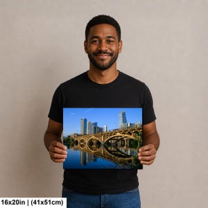 May include: A rectangular wall art print measuring 16 by 20 inches, featuring a scenic view of a concrete arched bridge spanning a calm river with a city skyline in the background under a clear blue sky, reflecting the structure in the water.