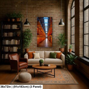 May include: A three-panel wall art print depicting a long, symmetrical corridor with orange-lit shelving units and a blue-toned ceiling. The artwork measures 36 by 72 inches and is displayed on a brick wall above a beige sofa in a living room.