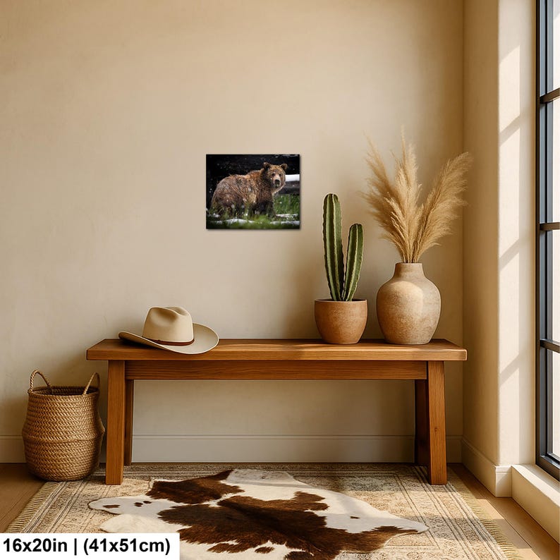May include: A rectangular wall art print featuring a brown bear standing in a grassy area with patches of snow. The print is displayed on a beige wall above a wooden bench, a beige hat, a potted cactus, and a vase with dried grass.