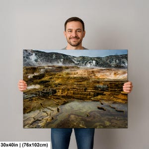 K&ouml;nnte beinhalten: Ein rechteckiger Wandkunstdruck in der Gr&ouml;&szlig;e 76 x 102 cm, der eine Landschaftsaufnahme von geothermischen Terrassen mit geschichteten Mineralablagerungen in Orange-, Braun- und Gelbt&ouml;nen zeigt, mit Dampf vor einer felsigen Bergkulisse.