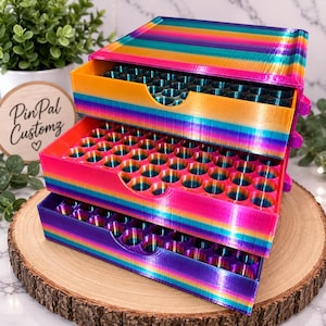 May include: A three-tiered storage organizer featuring a vibrant rainbow gradient finish. Each of the three stacked drawers contains a grid of circular cutouts designed for holding small items. The exterior displays horizontal stripes of orange, pink, blue, and purple.