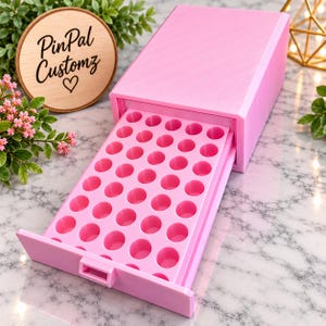May include: A light pink plastic storage organizer featuring a pull-out drawer with a grid of forty circular indentations for holding small items. The rectangular unit has a smooth finish and a small handle on the front of the drawer.