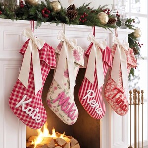 Puede incluir: Cuatro calcetines navideños colgados de una repisa de chimenea blanca. Los calcetines presentan patrones de cuadros rojos y blancos, motivos florales y rayas rojas y blancas. Cada calcetín está personalizado con los nombres bordados Kate, Miki, Rena y Tina.