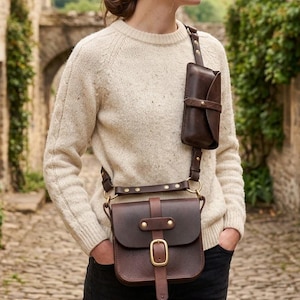 Puede incluir: Un bolso bandolera de cuero marrón oscuro con un compartimento principal rectangular que cuenta con un cierre de hebilla de latón y una bolsa vertical más pequeña unida a la correa, ambos asegurados con remaches y correas ajustables.