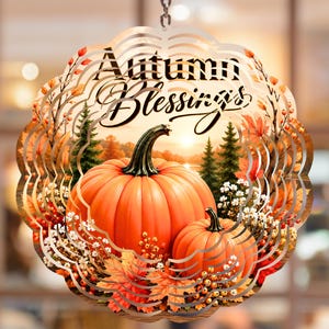 May include: A circular metal wind spinner featuring a printed autumn scene with two orange pumpkins, pine trees, and fall foliage. The text "Autumn Blessings" is printed in a black script font at the top of the scalloped-edge decorative hanging ornament.