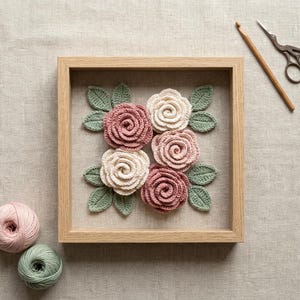 May include: A square light wood frame containing five crocheted roses in shades of dusty pink and cream, accented with crocheted sage green leaves. Beside the frame are two balls of yarn, a wooden crochet hook, and a pair of metal scissors.