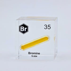 May include: A clear acrylic cube containing a sealed glass ampoule filled with a vibrant yellow liquid. The front face features the black chemical symbol Br inside a hexagon, the atomic number 35, the word Bromine, and the atomic weight 79.904.