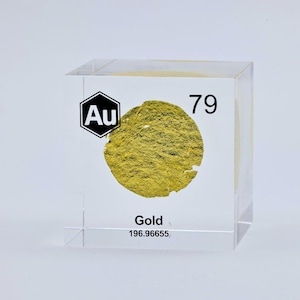 May include: A clear acrylic cube containing a textured, circular gold-colored sample. The front face features the chemical symbol Au inside a hexagon, the atomic number 79, the word Gold, and the atomic mass number 196.96655 printed in black.