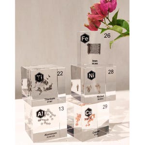 Puede incluir: Cuatro cubos de acrílico transparente que contienen muestras metálicas de hierro, níquel, cobre, titanio y aluminio. Cada cubo muestra el símbolo químico, número atómico, nombre y masa atómica. El cubo de hierro está sobre el de níquel.
