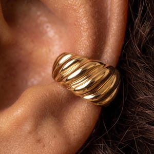 May include: A gold-toned ear cuff featuring a thick, polished, and ribbed croissant-style design. The jewellery piece has a rounded, chunky silhouette with multiple diagonal ridges that catch the light, creating a reflective and smooth metallic surface.