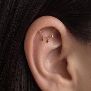May include: A gold ear cartilage chain featuring three small, dangling, teardrop-shaped charms. The delicate chain connects two points along the upper ear rim, allowing the three polished gold pendants to hang freely against the skin.