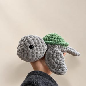 May include: A handmade crochet turtle plush toy featuring a soft grey yarn body and a textured green shell. The turtle has a rounded head with a single visible black plastic safety eye and four flippers extending from the main body.