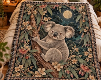 May include: A woven throw blanket featuring a central koala perched on a tree branch, surrounded by eucalyptus leaves and white and pink blossoms against a dark night sky with a full moon. The blanket has a decorative fringed edge.