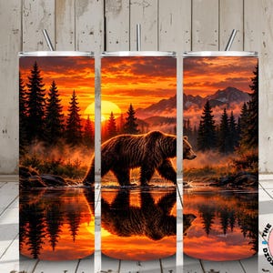 Könnte beinhalten: Ein zylindrischer Becher mit einem umlaufenden Motiv eines Braunbären, der bei Sonnenuntergang durch flaches Wasser läuft. Die Szene zeigt einen dichten Nadelwald, ferne Berge und eine leuchtend orangefarbene Sonne, die sich im Wasser spiegelt.