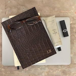 May include: A dark brown crocodile-textured notebook with the gold initials LR, resting on a light gray laptop. Included are brown-rimmed reading glasses, a beige Salt & Stone body lotion tube, and a black Dior lipstick with a silver CD logo.