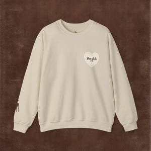 Peut inclure: Un sweat-shirt à col rond de couleur crème avec un petit motif en forme de cœur sur la poitrine gauche contenant le texte "Doe club". La manche droite présente une petite silhouette de cerf marron foncé et l'encolure porte une icône de cerf.