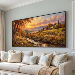 Op de afbeelding: Een rechthoekig landschapsschilderij in een zwarte lijst met een zonsondergang boven een glooiend landschap. De scène toont een stromende beek, een stenen brug, een klein huis op een heuvel en bomen met levendig oranje en geel herfstblad.