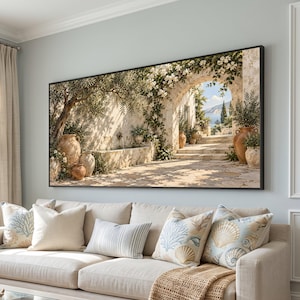 Op de afbeelding: Een rechthoekige wandkunstprint in een zwarte lijst met een mediterrane stenen binnenplaats met terracotta potten, klimmende witte bloemen en een stenen boogdeur die uitkijkt op de zee onder een blauwe lucht.
