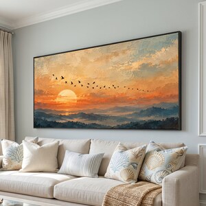 Könnte beinhalten: Ein großes rechteckiges Landschaftsgemälde in einem schwarzen Rahmen, das einen Sonnenuntergang über einem Gebirge zeigt. Der Himmel ist mit orangefarbenen und gelben Wolken sowie einem Vogelschwarm gefüllt. Im Vordergrund sind blaue Hügel.