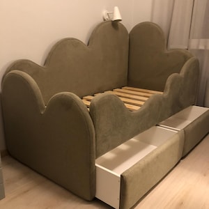 Op de afbeelding: Een saliegroen gestoffeerd bedframe met een geschulpt, wolkachtig hoofdeinde en zijpanelen. Het bed is voorzien van een houten lattenbodem en twee witte uitschuifbare opberglades aan de onderkant van het frame voor extra opbergruimte.