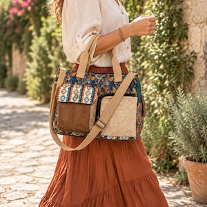 Puede incluir: Un bolso tote con múltiples patrones, base floral azul oscuro, dos bolsillos frontales de ante marrón y tela beige texturizada, una correa de lona beige y un asa superior. El bolso muestra un borde geométrico colorido en la solapa.