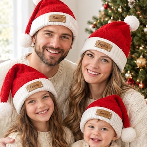 May include: Four red and white knit Santa hats featuring a white pom-pom at the top and a tan leather patch on the front cuff. Each patch is engraved with a name: David, Sophia, Emma, or Lucas. The hats have a textured cable knit pattern.