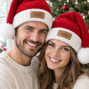 May include: Two red and white knit Santa hats featuring a white pom-pom at the tip and a brown leather patch on the front cuff. The left hat is engraved with the name David, and the right hat is engraved with the name Sophia.