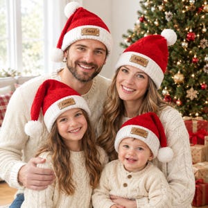 May include: Four red and white knit Santa hats featuring a white pom-pom at the top and a tan leather patch on the front cuff. Each patch is engraved with a name: David, Sophia, Emma, or Lucas. The hats have a textured cable knit pattern.