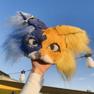 Può includere: Una maschera felina bicolore con il lato sinistro blu scuro con pelo lungo blu e il lato destro giallo con pelo lungo giallo. La maschera presenta dettagli oculari dipinti, un muso bianco, un piccolo ornamento a forma di osso e un simbolo solare.