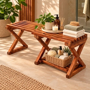 May include: A brown wooden bench with an X-shaped leg design and a slatted top. The bench features a lower shelf holding a woven wicker basket filled with bath accessories, while the top surface holds a potted plant, candle, and folded towels.
