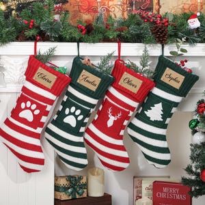Puede incluir: Cuatro calcetines navideños de punto colgados en fila, con rayas alternas rojas y blancas o verdes y blancas. Cada calcetín tiene una etiqueta con nombre en imitación cuero y un aplique de fieltro blanco con forma de huella, ciervo o árbol.