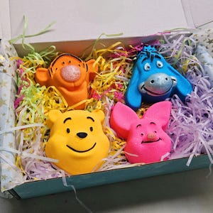 May include: A set of four character-shaped bath bombs in a box filled with colourful paper shreds. The bath bombs include an orange tiger, a blue donkey, a yellow bear, and a pink pig, each featuring distinct facial features and expressions.