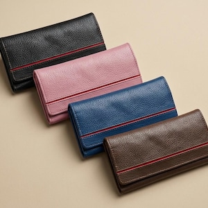 Peut inclure: Quatre portefeuilles rectangulaires à texture grainée, disposés en diagonale. Chaque portefeuille présente une bande décorative rouge horizontale sur le rabat avant. Les couleurs incluent noir, rose clair, bleu roi et marron foncé.
