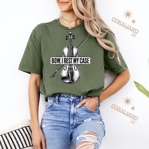 May include: An olive green short-sleeve t-shirt featuring a black and white graphic of a violin and a bow. The text "BOW, I REST MY CASE" is printed across the centre of the violin. The shirt is paired with light blue distressed denim jeans.