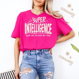 Op de afbeelding: Een felroze t-shirt met korte mouwen en de witte tekst SUPER INTELLIGENCE en de kleinere tekst (BEFORE 'SUPER INTELLIGENCE' WAS A THING) op de borst gedrukt. Het t-shirt wordt gecombineerd met een lichtblauwe spijkerbroek met scheuren.