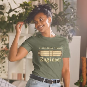 May include: An olive green t-shirt featuring the text "classically trained" above an illustration of a slide rule and the word "Engineer" below it in a light-coloured, vintage-style font. The shirt has a crew neckline and short sleeves.